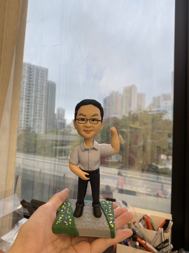 老闆主題的人像公仔立於灰色步道與綠地花卉點綴的底座上,由手掌托起展示。公仔為短髮、配戴眼鏡,穿淺灰短袖襯衫、黑色長褲與黑色皮鞋;右臂上舉作打氣姿勢,左手自然放置。背景為臨窗的辦公環境,可見城市高樓與道路景觀,前景擺有文具收納盒。