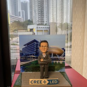 一位男性人像公仔站在石磚步道造型底座中央，雙手交叉、身穿深灰色西裝與條紋襯衫，配戴眼鏡，神情自信。背板為現代科技園區或創新中心風格的建築與水景照片。底座前方有藍白色牌匾，印有「CREE LED」字樣與箭頭圖示。整體呈現科技與照明產業氛圍的人像公仔展示。