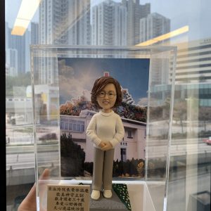 透明展示盒中的院長榮休主題人像公仔：短捲髮、戴圓框眼鏡的女性公仔，穿白色麻花針織上衣、米色長褲與白鞋，雙手交疊站立於方形底座。身後背板為教堂建築照片，屋頂有紅色十字架，周圍樹木花海；底座一角鋪有綠草與小白花。展示盒左前方立有中文感謝銘牌，提及榮休賀辭與「神召會聖光堂」。整體由手托起，背景是窗外城市高樓景色。