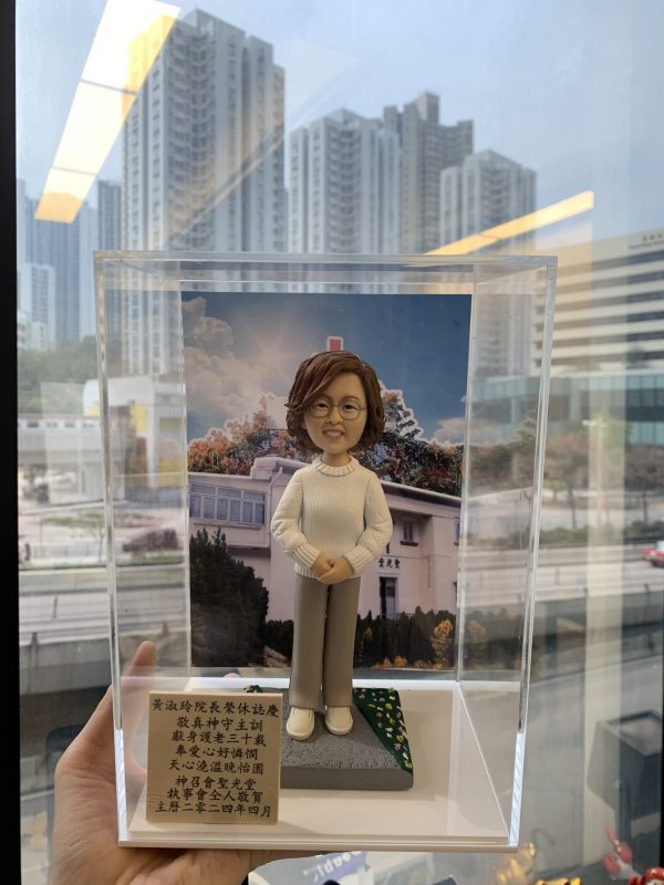 透明展示盒中的院長榮休主題人像公仔：短捲髮、戴圓框眼鏡的女性公仔，穿白色麻花針織上衣、米色長褲與白鞋，雙手交疊站立於方形底座。身後背板為教堂建築照片，屋頂有紅色十字架，周圍樹木花海；底座一角鋪有綠草與小白花。展示盒左前方立有中文感謝銘牌，提及榮休賀辭與「神召會聖光堂」。整體由手托起，背景是窗外城市高樓景色。