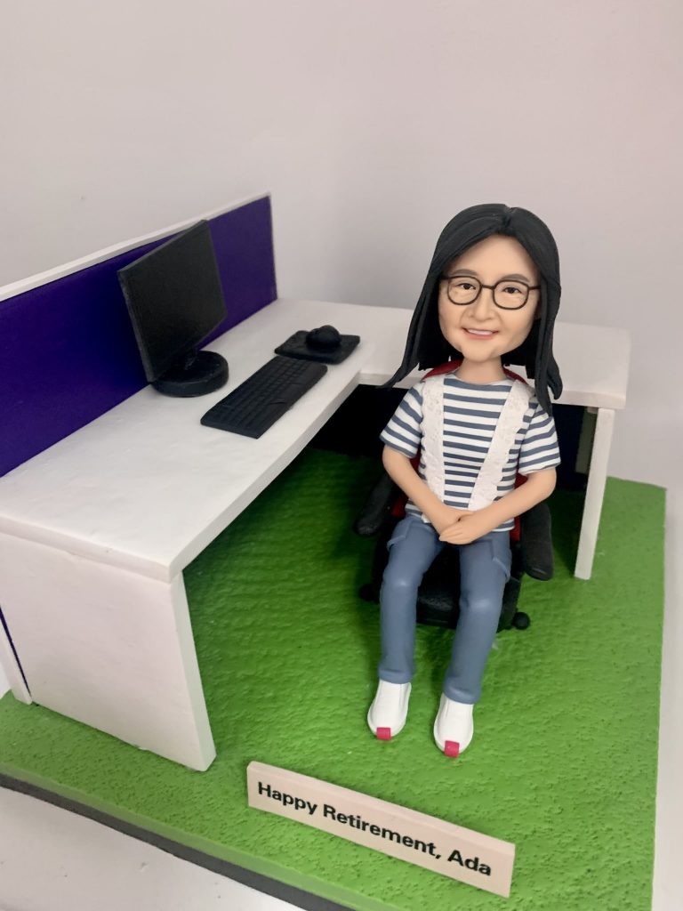 一款女性人像公仔坐在黑色辦公椅上，穿藍白條紋上衣、淺藍長褲與白色鞋，配戴眼鏡並微笑。旁邊為白色L形辦公桌與紫色桌屏，桌上擺有黑色螢幕、鍵盤、滑鼠與滑鼠墊。地台為草綠色質感，前方木質名牌刻有退休祝賀字樣。