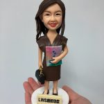 一款女性人像公仔被手掌托起展示。公仔留長深棕髮、微笑，身穿深棕色V領連身裙與黑色平底鞋；右臂抱著粉紅到藍綠漸層封面的冊子，左手提黑色手袋。站在白色圓形底座上，前方有淺米色名牌刻字「LWS創校功臣」。背景為素灰白色。