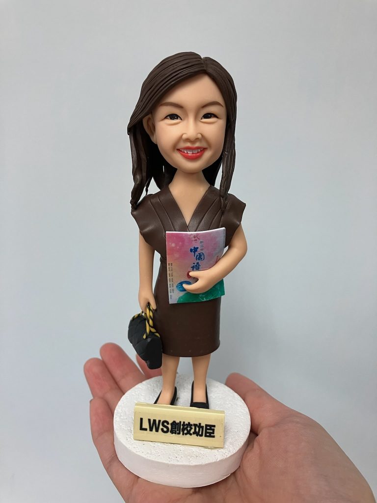 一款女性人像公仔被手掌托起展示。公仔留長深棕髮、微笑，身穿深棕色V領連身裙與黑色平底鞋；右臂抱著粉紅到藍綠漸層封面的冊子，左手提黑色手袋。站在白色圓形底座上，前方有淺米色名牌刻字「LWS創校功臣」。背景為素灰白色。