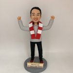 人像公仔立於圓形灰色底座上，雙手高舉呈慶祝姿勢；短髮、面帶笑容，穿灰色連帽上衣、黑色長褲與紅白運動鞋，頸上繫紅白相間條紋圍巾。底座前方金色銘牌印有英文字：「Steven Lee / 18 Years with AQUA!」。