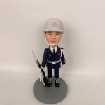 穿深藍制服與白色安全帽的男性人像公仔，戴白色頤帶與肩繩，胸前有彩色識別條與白色腰帶；右手持黑白配色禮槍，黑靴踏立於灰色圓形底座。背景為純白牆面。