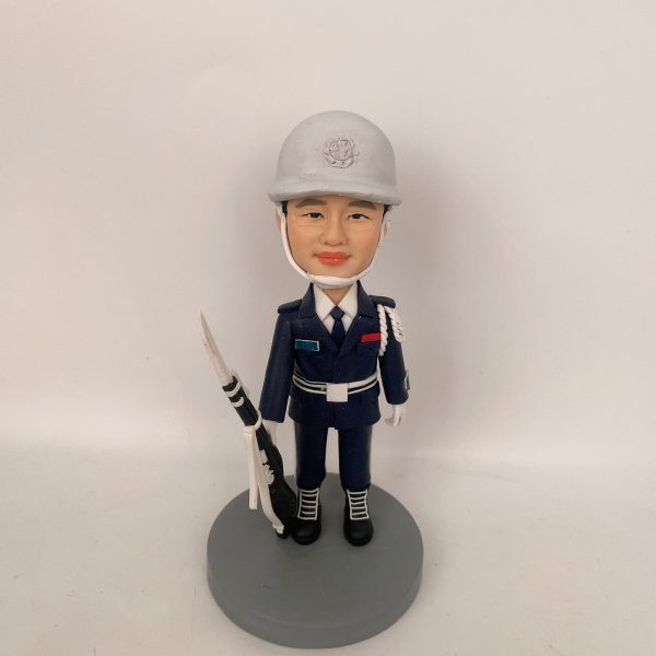 穿深藍制服與白色安全帽的男性人像公仔，戴白色頤帶與肩繩，胸前有彩色識別條與白色腰帶；右手持黑白配色禮槍，黑靴踏立於灰色圓形底座。背景為純白牆面。