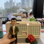 男性人像公仔穿白襯衫與綠色外套、黑長褲與黑鞋，雙手於身前交握，站在灰色情境底座上。背後是帶有「Housing Authority Headquarters」與「房屋委員會」標示的白色建築模型，右前方有紅色幾何雕塑與綠色草叢。底座前方立有白色名牌寫著「Happy Retirement to Kenneth」。畫面邊緣可見工作檯與城市窗景。