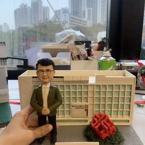 男性人像公仔穿白襯衫與綠色外套、黑長褲與黑鞋，雙手於身前交握，站在灰色情境底座上。背後是帶有「Housing Authority Headquarters」與「房屋委員會」標示的白色建築模型，右前方有紅色幾何雕塑與綠色草叢。底座前方立有白色名牌寫著「Happy Retirement to Kenneth」。畫面邊緣可見工作檯與城市窗景。