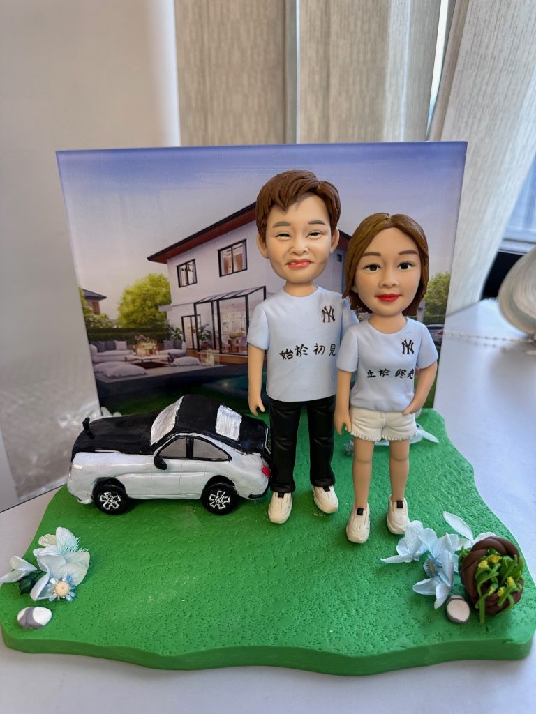 草綠不規則底座上的情侶人像公仔：左側男性穿淺藍T恤與黑長褲、白色運動鞋；右側女性穿同色系T恤搭白色短褲與白鞋。兩人並肩站立，身後擺放黑白配色的私家車模型；背景立起一幅現代獨棟住宅庭院的畫布，看似作為情境背板。底座兩側有藍白花材與小石頭、右前方放有裝綠葉的棕色小籃。