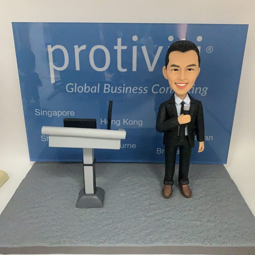 商務演講主題的人像公仔：一位短髮男性穿黑色西裝、白襯衫與領帶，右手持麥克風微笑站立；左側是灰白配色的講台，上面擺放筆電與設備。背景背板印有「protiviti – Global Business Consulting」與多個城市名稱（如 Singapore、Hong Kong 等），地台為灰色細紋質感。整體呈現顧問公司於會議/培訓中的專業講者場景的人像公仔。