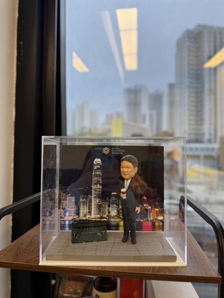 一款人像公仔置於透明展示盒中,背景為維多利亞港夜景與高樓天際線;地台為灰色石板路紋理。公仔穿深色西裝、白襯衫與條紋領帶,右手拿外帶咖啡杯,站姿輕鬆。腳邊擺放一台深綠色雙層電車模型;背景板可見英文字樣與標誌。整體放在木層板上,窗外是城市高樓景致。
