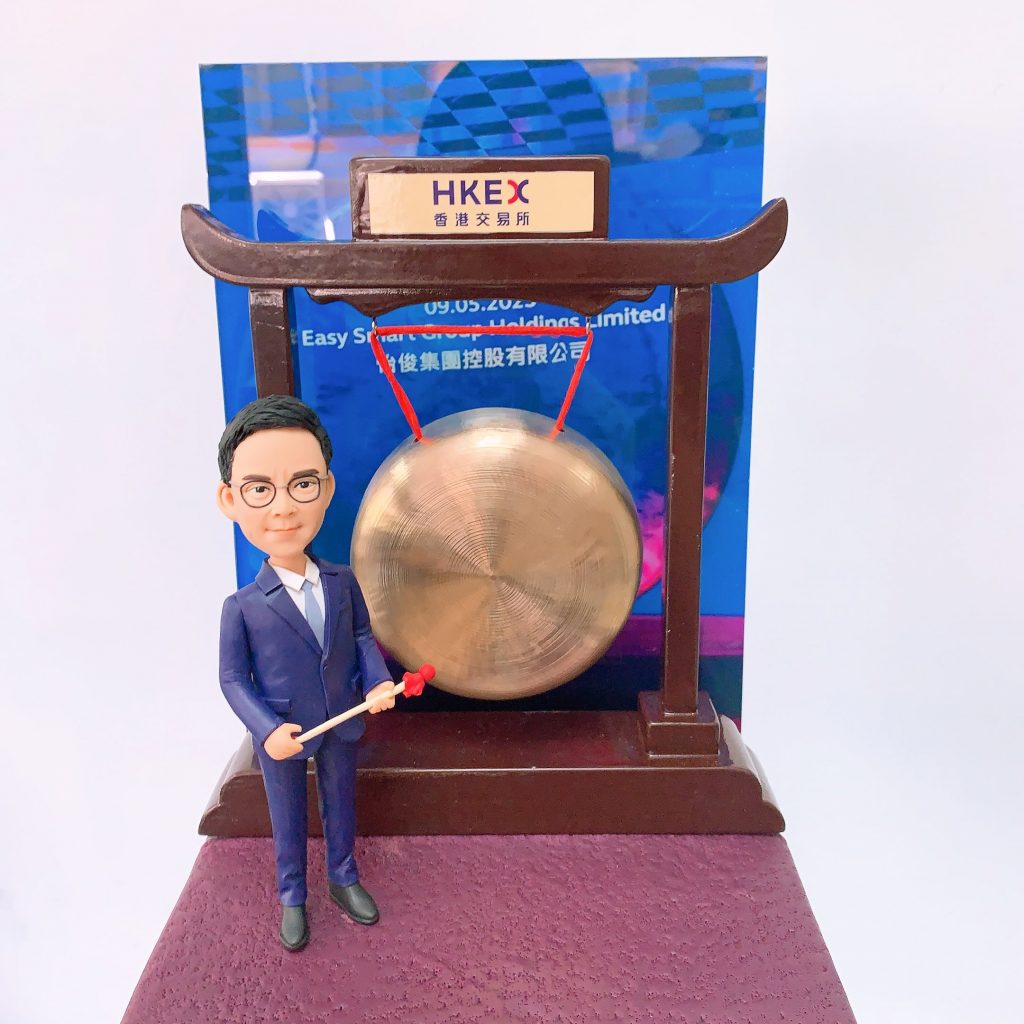 以香港交易所敲鑼儀式為主題的人像公仔：身穿深藍西裝與白襯衫的男性公仔，手持紅頭木槌站在銅鑼旁；銅鑼懸掛於深木色拱形架上，頂部銘牌印有「HKEX 香港交易所」。背板為藍色活動海報樣式，底座為暗紫色質感平台。整體呈現企業代表在港交所參與上市敲鑼的慶賀場景，作為金融領域的紀念人像公仔。