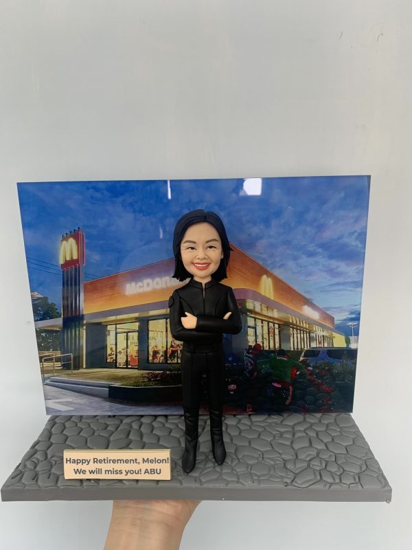 退休致敬主題的人像公仔：黑色長髮女性，身穿全黑制服外套與長褲、黑色長靴，雙手交叉自信站姿，面帶微笑。腳下為灰色石板紋地台，前方有英文字牌：「Happy Retirement, Melon! We will miss you! ABU」。背板為夜色中的 McDonald’s 麥當勞門店外觀，招牌金色拱門清晰可見。整體呈現餐飲門店管理人員的榮休祝福禮物場景。