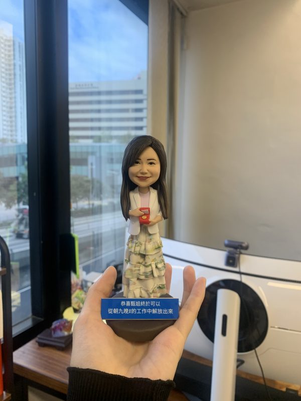 同事退休主題的人像公仔：女同事長髮微笑，穿白色外套與多層荷葉邊長裙，手持紅色禮盒站立；深藍色斜角名牌座刻有中文祝福文字。背景為辦公室窗景與城市大樓，整體呈現精緻小巧、可單手托起的桌上型退休禮物的人像公仔。