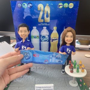 飲品品牌20週年主題的人像公仔場景：一男一女公仔穿著藍色K2品牌T恤與球鞋，微笑站立，雙手共同舉著藍色宣傳橫幅（印有K2與NO.1字樣）；右側白色圓桌擺放多款彩色瓶裝飲品模型。背板是藍底「20th LIFE WATER 20th ANNIVERSARY」設計，並有多款瓶裝水與飲料圖像；地台為灰色地磚，前方小牌寫有“Congratulations! From: Hong Kong Cousin (2023.8.13)”。整體呈現慶祝品牌里程碑的紀念公仔擺設。