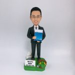 穿深灰色西裝、打條紋領帶的男性人像公仔，戴眼鏡、微笑站立，雙手於身前捧著一本藍色封面的文件冊。公仔置於草綠色地台，腳邊有白色指示牌造型與小型告示牌（印有「最佳 OPPA 最高老闆 JFIU SEC 4 以上」字樣），右側點綴麻繩圈與黃色小花及銀色小物件。背景為純白色，整體呈現尊敬老闆與退休致意的禮贈氛圍。