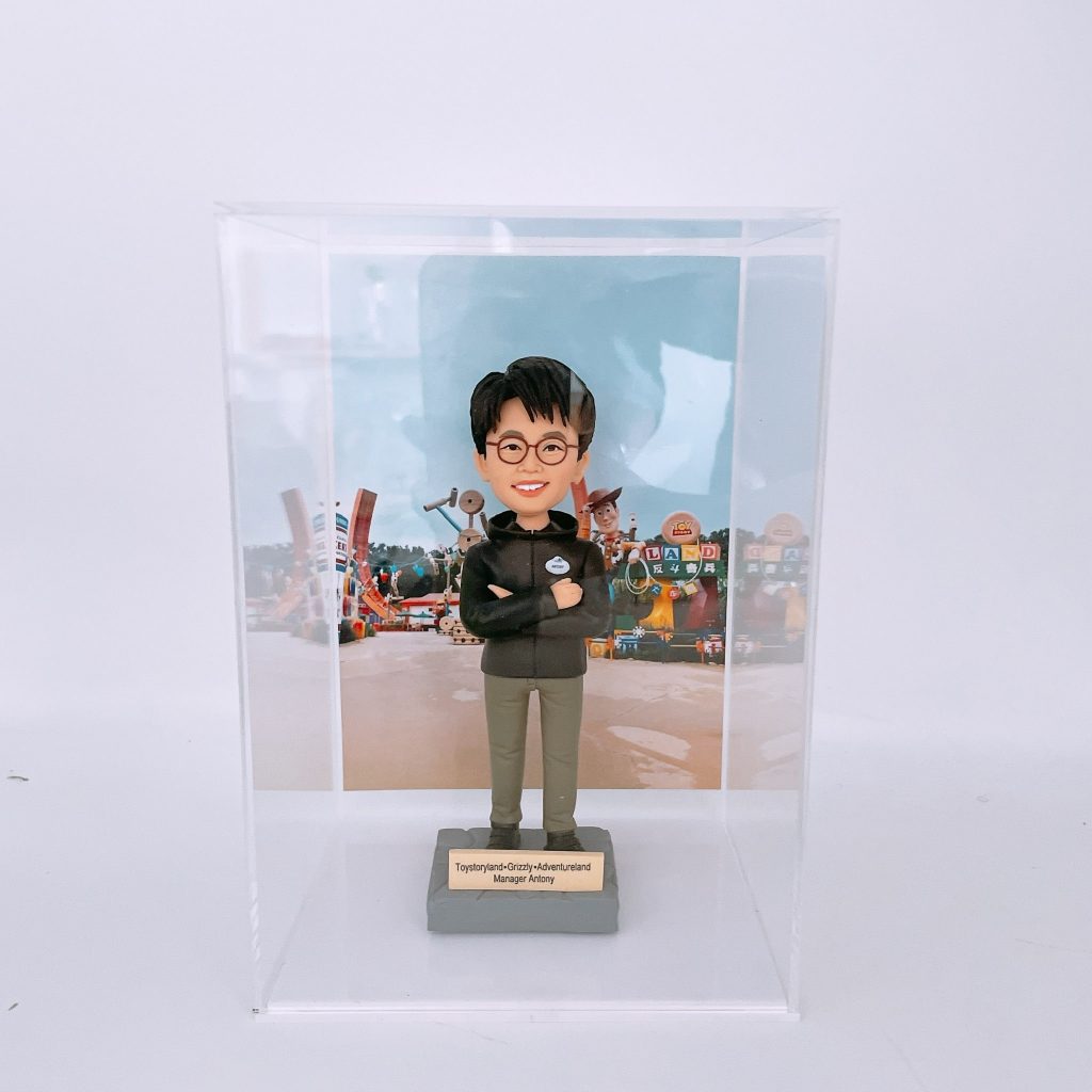 一款人像公仔置於透明展示盒內，背景是色彩繽紛的主題樂園布景（玩具造型與字母積木裝飾）。公仔戴黑框眼鏡，穿深色連帽外套與卡其長褲、黑白運動鞋，胸前配有圓形名牌，雙臂交叉站立在灰色底座上。底座金屬牌刻有「Toystoryland • Grizzly • Adventureland Manager Antony」等字樣。
