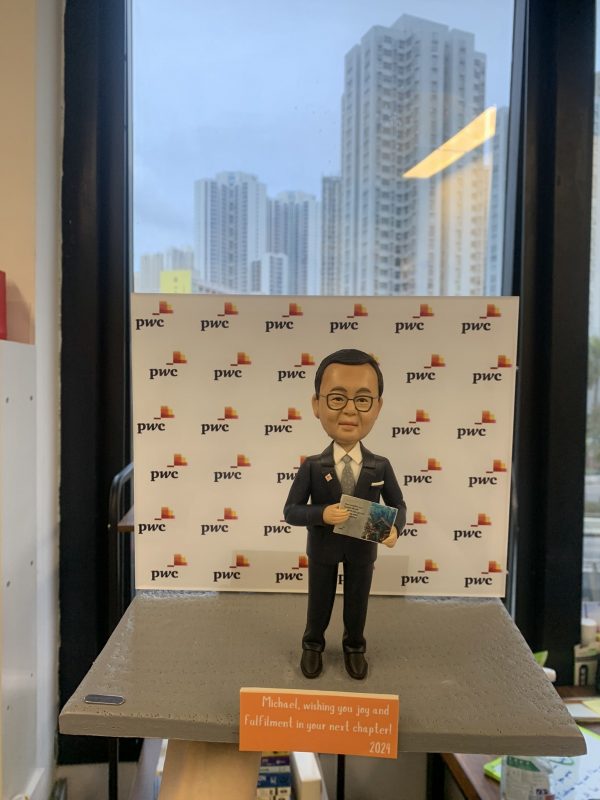 人像公仔站在灰色台座上，身穿深色西裝與領帶，配戴眼鏡，手持一本印有城市景觀圖的冊子；背板重複出現「pwc」與彩色方塊標誌，前緣橘色小牌寫有祝福語與年份2024；場景位於室內窗前，可見遠處高樓天際線。整體呈現專業商務與顧問氛圍的人像公仔展示。