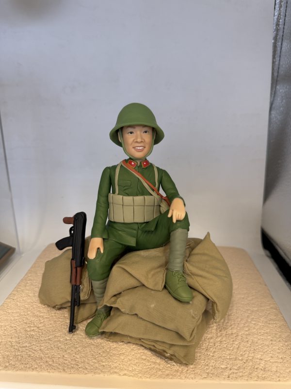 一款軍事風格的人像公仔坐在米色沙包堆上，身穿綠色軍裝與同色頭盔，胸前配有彈藥攜行具與斜揹背帶，領口見紅色領章細節；左側倚著黑棕色步槍道具。公仔面帶微笑，右膝抬起、左腿下垂，背景為淺色展示空間。