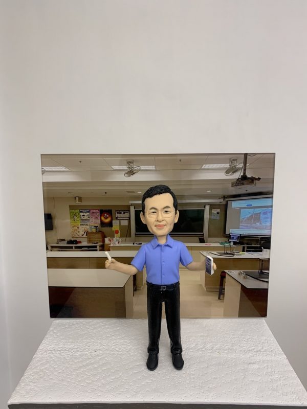 教室主題的人像公仔：短髮男性公仔身穿藍色短袖襯衫與黑色長褲，雙臂微張站立，右手持白色粉筆、左手拿書本。公仔置於顆粒感白色台座前，背板為教室場景，包含黑板、投影幕、講桌、實驗桌與牆上海報。