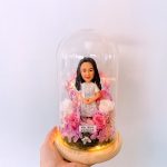 玻璃罩永生花擺設中的人像公仔：長黑髮女性公仔穿淺灰白蕾絲連衣裙與白色小外套，雙手交握於身前，站在圓形木質底座上。罩內以粉紅、玫瑰粉與白色的永生玫瑰與繡球花環繞，底部鋪苔蘚並點綴暖黃燈串。前方有一條白色小字牌，寫有中文祝福語。整體由手掌托起，背景為純白牆面。