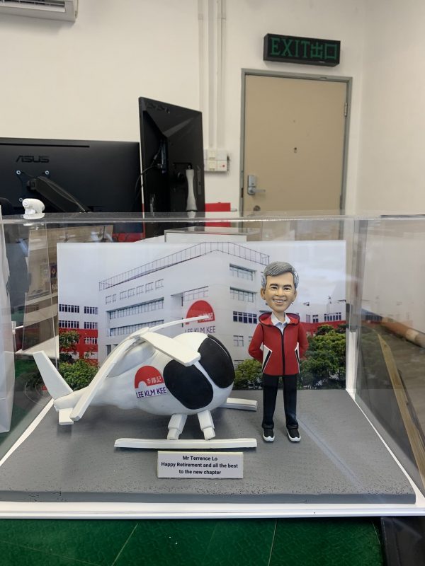 退休祝福主題的人像公仔場景：一位短灰髮、微笑的男性身穿紅色連帽外套、黑長褲與黑色運動鞋，雙手插袋站立；旁邊是一架白色直升機模型，機身與艙門上印有紅色圓形圖案與「LEE KUM KEE」字樣。兩者置於灰色地台，前方有英文銘牌：「Mr Terrence Lo — Happy Retirement and all the best to the new chapter」。背景背板為公司工廠大樓外觀與品牌標誌。整組作品收納在透明展示盒內，拍攝環境為室內辦公區與EXIT指示牌可見。整體呈現企業退休致意的人像公仔禮物。