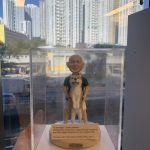 祝福主題的人像公仔：一位光頭微笑的男性穿黑色上衣與米色短褲，雙手在前方輕扶一隻站立的大型犬（米白毛色、粉鼻、吐舌神情），呈現親密互動。作品置於圓角米色底座，前側貼有英文字牌，內容為「To Dear Boss - Jeremy Masters ... Wishing ‘Boss’ all the best ... Sincerely, OPs Team (Grace, Phyllis, Wing, Joy)」等祝福語。整體收納在透明防塵展示盒中，由手掌承托；背景為高樓林立的城市窗景與晴朗藍天。整體氛圍溫暖，適合作為老闆或親友的祝福禮物人像公仔。