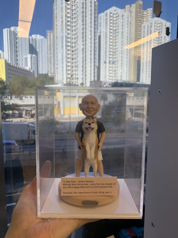 祝福主題的人像公仔：一位光頭微笑的男性穿黑色上衣與米色短褲，雙手在前方輕扶一隻站立的大型犬（米白毛色、粉鼻、吐舌神情），呈現親密互動。作品置於圓角米色底座，前側貼有英文字牌，內容為「To Dear Boss - Jeremy Masters ... Wishing ‘Boss’ all the best ... Sincerely, OPs Team (Grace, Phyllis, Wing, Joy)」等祝福語。整體收納在透明防塵展示盒中，由手掌承托；背景為高樓林立的城市窗景與晴朗藍天。整體氛圍溫暖，適合作為老闆或親友的祝福禮物人像公仔。