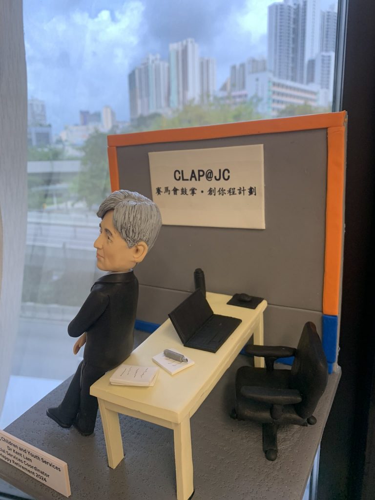 退休致敬主題的人像公仔：銀灰短髮男性穿黑色西裝與條紋領帶，側身倚靠米色長桌；桌上有開啟的筆電、筆與文件本、紙張疊。背板印有「CLAP@JC」與中文標語，台座前方附有英文名牌，標註社會服務相關職稱與退休祝福。整體置於室內窗邊場景中，可見城市高樓與藍天背景，呈現社福教育機構辦公氛圍的人像公仔。