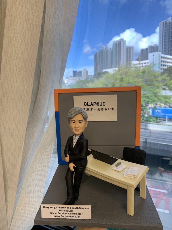 退休致敬主題的人像公仔：銀灰短髮男性穿黑色西裝與條紋領帶，側身倚靠米色長桌；桌上有開啟的筆電、筆與文件本、紙張疊。背板印有「CLAP@JC」與中文標語，台座前方附有英文名牌，標註社會服務相關職稱與退休祝福。整體置於室內窗邊場景中，可見城市高樓與藍天背景，呈現社福教育機構辦公氛圍的人像公仔。