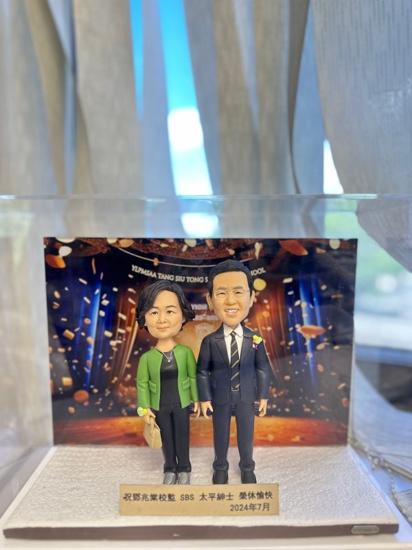 兩款並立的人像公仔置於白色砂岩質感底座上，背板為舞台與金色彩帶燈光效果。左側女性公仔留短捲髮，穿綠色外套配黑色上衣與長褲、灰色鞋，右手持米色手袋；右側男性公仔戴眼鏡、穿黑色西裝與白襯衫、黑黃條紋領帶，胸前別花。前方金色名牌刻有中文祝賀字樣，內容包含「祝賀××校監…榮休愉快 2024年7月」等。整體呈現典禮與賀壽氣氛。