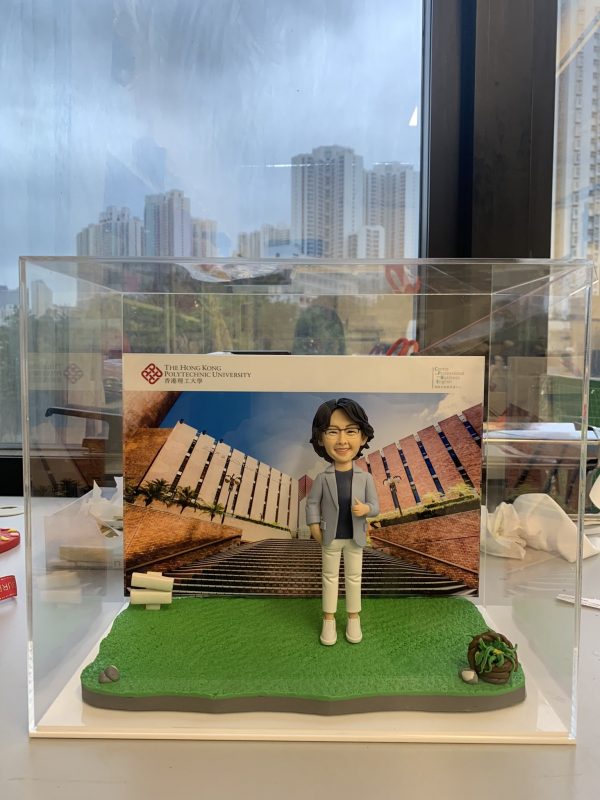 透明壓克力展示盒內，一尊短髮戴眼鏡的人像公仔站在綠色草地質感底座上，穿淺灰外套、深色上衣、白色九分褲與白鞋，右手比讚、左手插口袋。背板是一張校園建築階梯的照片，左上角印有「The Hong Kong Polytechnic University／香港理工大學」及紅色結形校徽，右上角標示「College of Professional and ...」等英文字樣。底座右前角有一小叢多肉植物與石頭裝飾；盒外背景透出城市高樓景色。
