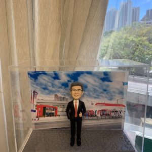 戶外企業場景的人像公仔:中年男性穿黑色西裝與紅色領帶,戴眼鏡微笑比讚,站在灰色石板路面底座上;背板為中油站/加油站樣式的大型場景,可見紅白建築、加油島與車道,以及藍天白雲與城市高樓天際線。整體置於透明展示盒內,呈現能源/石油零售產業形象的人像公仔。