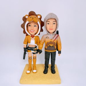 一對情侶主題人像公仔並肩站在淺黃色方形底座上：左側女性穿獅子頭造型連帽與棕色鬃毛、黃橘色短版上衣與短褲，腿上貼有獅子貼布，腳穿長筒襪與球鞋，腰配皮帶與小刀具道具；右側男性著灰色連帽外套，胸前有獅子圖樣，黑色長褲與靴子，背後背帶配件可見。兩人牽手，色調以黃橘與棕、灰為主，背景純白。