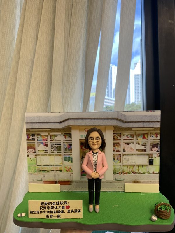 一款老師主題的人像公仔置於校園場景底座：公仔留短髮、戴眼鏡，穿粉色外套、印花上衣與黑色長褲，雙手於身前交握；腳踏灰色地磚，站在綠色草地底座上。背景板為幼稚園/兒童學校門口與窗格，窗內有彩繪與兒童圖案。前方立有中文祝福牌匾，內容祝賀校長退休；右側有小花籃與白色小裝飾物。整體陳列在室內、靠近落地窗與窗簾。