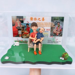 退休主題的人像公仔場景:短髮女性公仔穿紅色格紋上衣與黑裙,腳踩橘色鞋,坐在米色單人椅上懷抱笑臉嬰兒公仔;底座為草地造型,右前方有小花籃與石頭擺飾,左側有白色卷軸。背板為「榮休之喜」主題相片牆,包含多張生活照與感謝字句,呈現同事祝福、回顧服務年資與退休快樂氛圍的人像公仔。