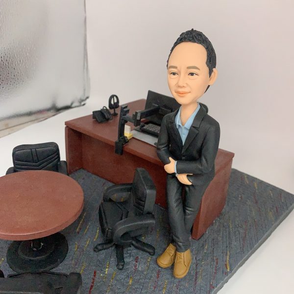 人像公仔辦公室場景：短髮男性公仔穿深色西裝外套與長褲、內搭淺藍襯衫，腳穿棕色皮靴，側身倚靠木質主管桌，雙手自然交疊。桌上配置筆電、螢幕支架、電話座與文具收納；一旁有黑色辦公椅與圓形會議桌，地面為帶有彩色線條的地毯紋理。整體呈現主管/經理級專業辦公氛圍的人像公仔擺設。