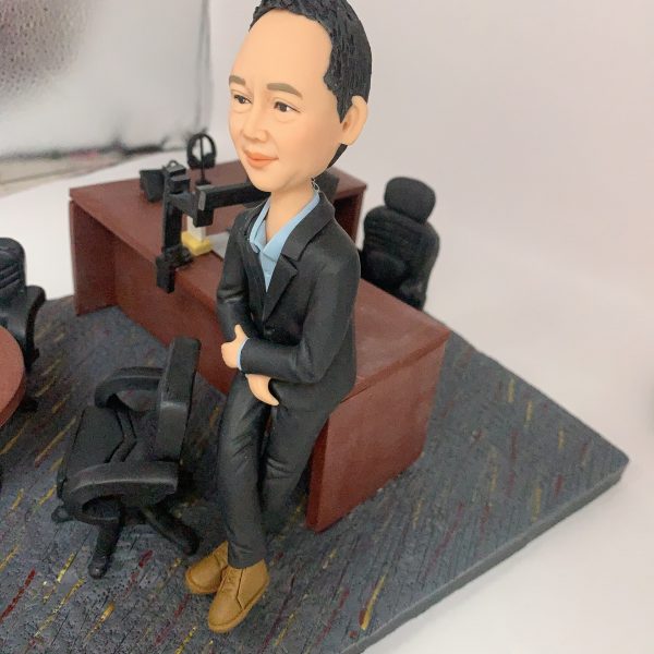 人像公仔辦公室場景：短髮男性公仔穿深色西裝外套與長褲、內搭淺藍襯衫，腳穿棕色皮靴，側身倚靠木質主管桌，雙手自然交疊。桌上配置筆電、螢幕支架、電話座與文具收納；一旁有黑色辦公椅與圓形會議桌，地面為帶有彩色線條的地毯紋理。整體呈現主管/經理級專業辦公氛圍的人像公仔擺設。