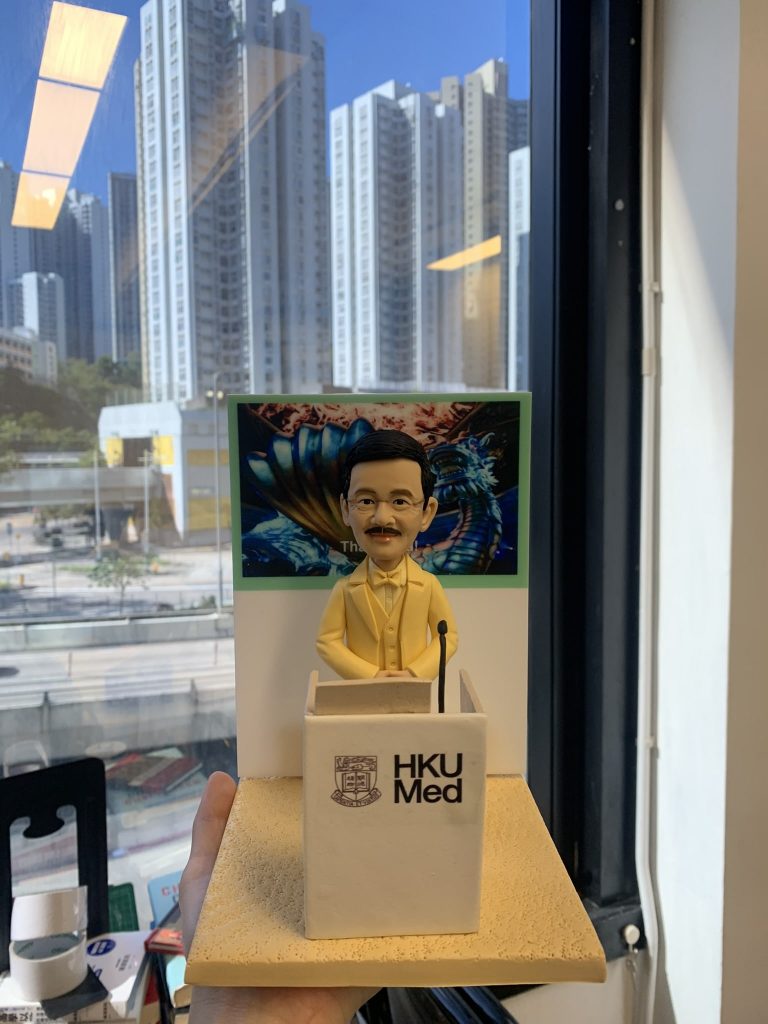 老師禮物人像公仔，人物戴眼鏡、留短鬚，身穿黃色西裝，站在印有「HKU Med」字樣的講台後方，雙手放在講台上，神態莊重。