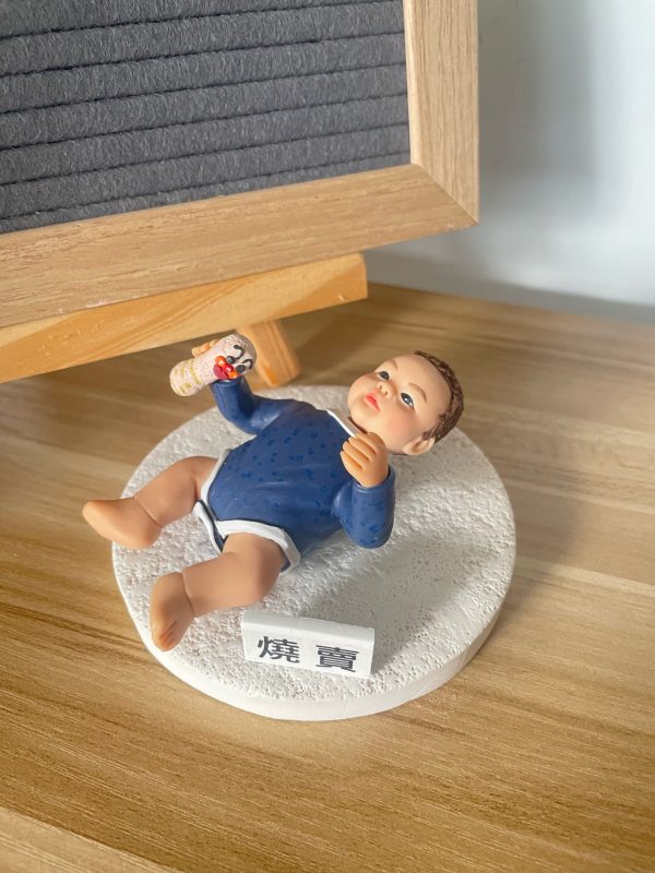 木紋桌面上擺放一款小朋友主題bb人像公仔：嬰兒造型側躺在白色圓形凹凸紋理底座上，穿深藍長袖上衣與白色尿布褲，右手握著奶瓶圖案的小道具，臉頰紅潤、表情放鬆；底座前方有小牌寫著「燒賓」。背景為木框黑灰字母板與牆面。