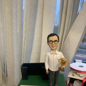 人像公仔獎勵主題場景：男性造型，短髮戴黑框眼鏡，穿白色長袖襯衫、深灰長褲與黑鞋，右手拿金色獎盃（杯身刻有功字），站在綠色紋理底座上。身後有黑色辦公沙發造型裝飾，前方放置附花飾的小牌，牌面文字表達對領導的尊敬。背景為垂直布簾與窗景，整體呈現表揚、感謝與企業榮耀氛圍的人像公仔。