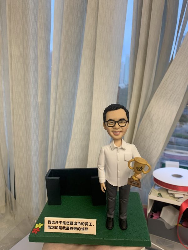 人像公仔獎勵主題場景：男性造型，短髮戴黑框眼鏡，穿白色長袖襯衫、深灰長褲與黑鞋，右手拿金色獎盃（杯身刻有功字），站在綠色紋理底座上。身後有黑色辦公沙發造型裝飾，前方放置附花飾的小牌，牌面文字表達對領導的尊敬。背景為垂直布簾與窗景，整體呈現表揚、感謝與企業榮耀氛圍的人像公仔。