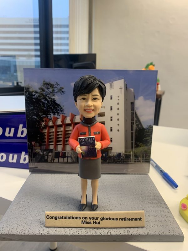 一款歷史老師主題的人像公仔站在灰色紋理底座上，身穿橙色上衣與深灰色半身裙，雙手捧著一本封面寫有「WORLD HISTORY」的書本。背後立有校園建築背景板，可見白色外牆與十字架標誌。底座前方有英文祝賀牌寫著「Congratulations on your glorious retirement Miss Hui」。桌面場景上還有藍色原子筆與辦公用品。