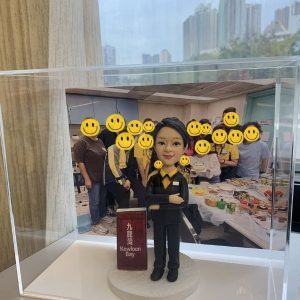 透明壓克力展示盒內,一個穿黑色西裝、黃色襯衫的人像公仔雙手交疊站立,腳下為白色圓形底座;旁邊立著寫有「九龍灣 Kowloon Bay」的紅色站牌造型。展示盒後方放置一張團體合照,十多位穿黃黑制服與便服的人在辦公室/茶水間前合影,桌面擺滿食物與飲品。窗外可見高樓天際線與陰天。