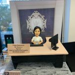 律師榮休主題的人像公仔：黑髮女律師公仔身穿白色上衣，端坐於淺膚色桌面、黑色桌腳的律師辦公桌後方，雙手置於鍵盤前。桌面右側擺放黑色顯示器與滑鼠，左前方立有金銅色名牌，刻有祝賀榮休與職銜資訊。背板為律師事務所徽章與字母標識的紫色底圖，前景底座呈現灰黑木紋質感。畫面在辦公室靠窗拍攝，可見窗外城市高樓景與室內書籍雜物，整體營造專業、莊重且具儀式感的律師榮休禮物與律師人像公仔。