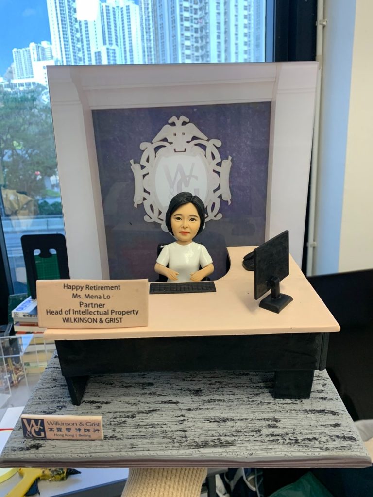 律師榮休主題的人像公仔：黑髮女律師公仔身穿白色上衣，端坐於淺膚色桌面、黑色桌腳的律師辦公桌後方，雙手置於鍵盤前。桌面右側擺放黑色顯示器與滑鼠，左前方立有金銅色名牌，刻有祝賀榮休與職銜資訊。背板為律師事務所徽章與字母標識的紫色底圖，前景底座呈現灰黑木紋質感。畫面在辦公室靠窗拍攝，可見窗外城市高樓景與室內書籍雜物，整體營造專業、莊重且具儀式感的律師榮休禮物與律師人像公仔。