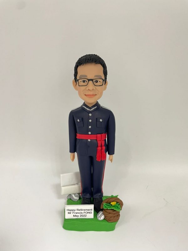 身穿深藍色制服的人像公仔直立於草地質感底座上；制服為立領、肩章與口袋翻蓋設計，胸前有金屬鏈與扣件，褲側有紅色側條，腰間束紅色流蘇腰帶，腳穿黑皮鞋；公仔短髮、戴黑框眼鏡、微笑。底座左後有白色告示牌與銀色配件，右前有編織籃盛放綠葉與黃色花；前置銘牌寫著 “Happy Retirement Mr Francis FONG May 2022”。