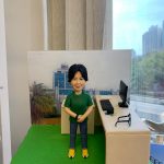 便衣風格的警察人像公仔：短黑髮人士穿綠色短袖上衣、深灰長褲與黃色運動鞋，雙手自然交握站立於綠色草地質感底座上；右側是一張米白色長桌，桌面擺放黑色螢幕、鍵盤、文件、鑰匙串與一副黑框眼鏡，呈現行政部人員常用的辦公配置。背板為藍白相間的現代化政府/警署建築外觀，象徵單位背景；左前角有圓形徽章樣式小裝飾。整體以室內靠窗拍攝，窗外可見藍天與高樓，風格清爽、專業，適合作為警務人員禮物或行政部人員的客製人像公仔。