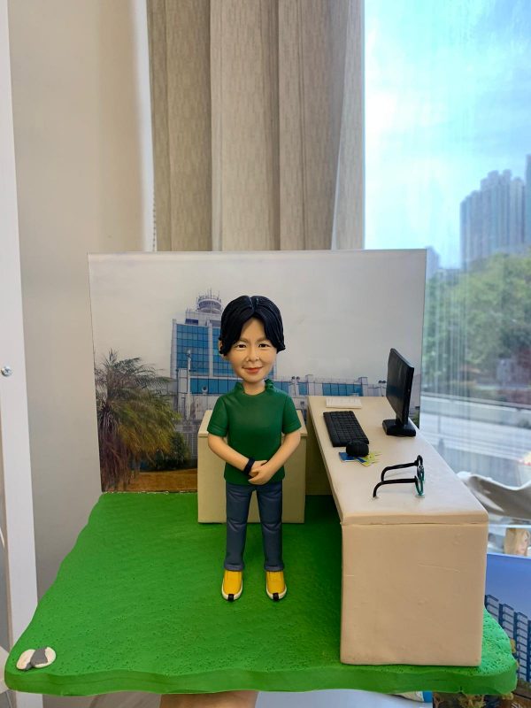 便衣風格的警察人像公仔：短黑髮人士穿綠色短袖上衣、深灰長褲與黃色運動鞋，雙手自然交握站立於綠色草地質感底座上；右側是一張米白色長桌，桌面擺放黑色螢幕、鍵盤、文件、鑰匙串與一副黑框眼鏡，呈現行政部人員常用的辦公配置。背板為藍白相間的現代化政府/警署建築外觀，象徵單位背景；左前角有圓形徽章樣式小裝飾。整體以室內靠窗拍攝，窗外可見藍天與高樓，風格清爽、專業，適合作為警務人員禮物或行政部人員的客製人像公仔。