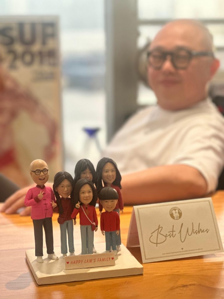 前景是一組家庭主題的人像公仔擺件：六位角色站在米色方形底座上，底座前有小牌寫「Happy Lam’s Family」。公仔皆穿紅色上衣與淺藍褲、白鞋，多人做出手指V手勢；其中左側一位戴黑框眼鏡、著粉紅外套。右前方擺放一張米白色祝賀卡，上書「Best Wishes」。背景為模糊的室內場景與坐著的成人身影。整體色調溫暖、桌面為木質。